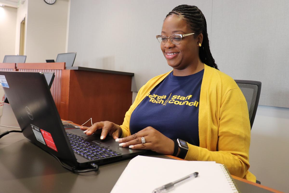 Woman in blue shirt and yellow sweater sits in front of laptop computer.